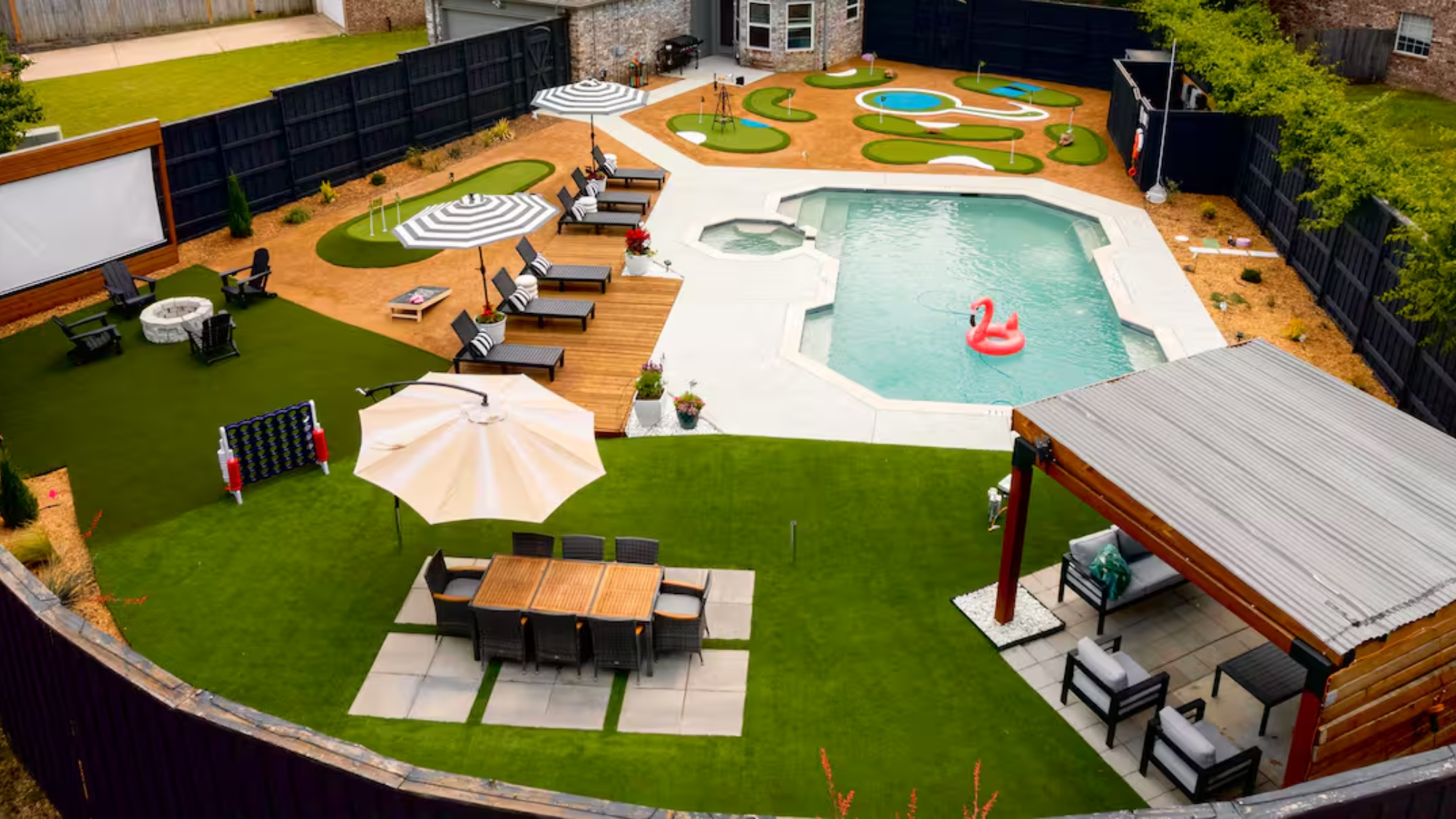 A family-friendly Frisco Airbnb with a spacious backyard featuring a swimming pool with a pink flamingo float, lounge chairs with striped umbrellas, a mini-golf area, an outdoor dining set under a large umbrella, a covered patio seating area, a fire pit with chairs, and an outdoor movie screen.