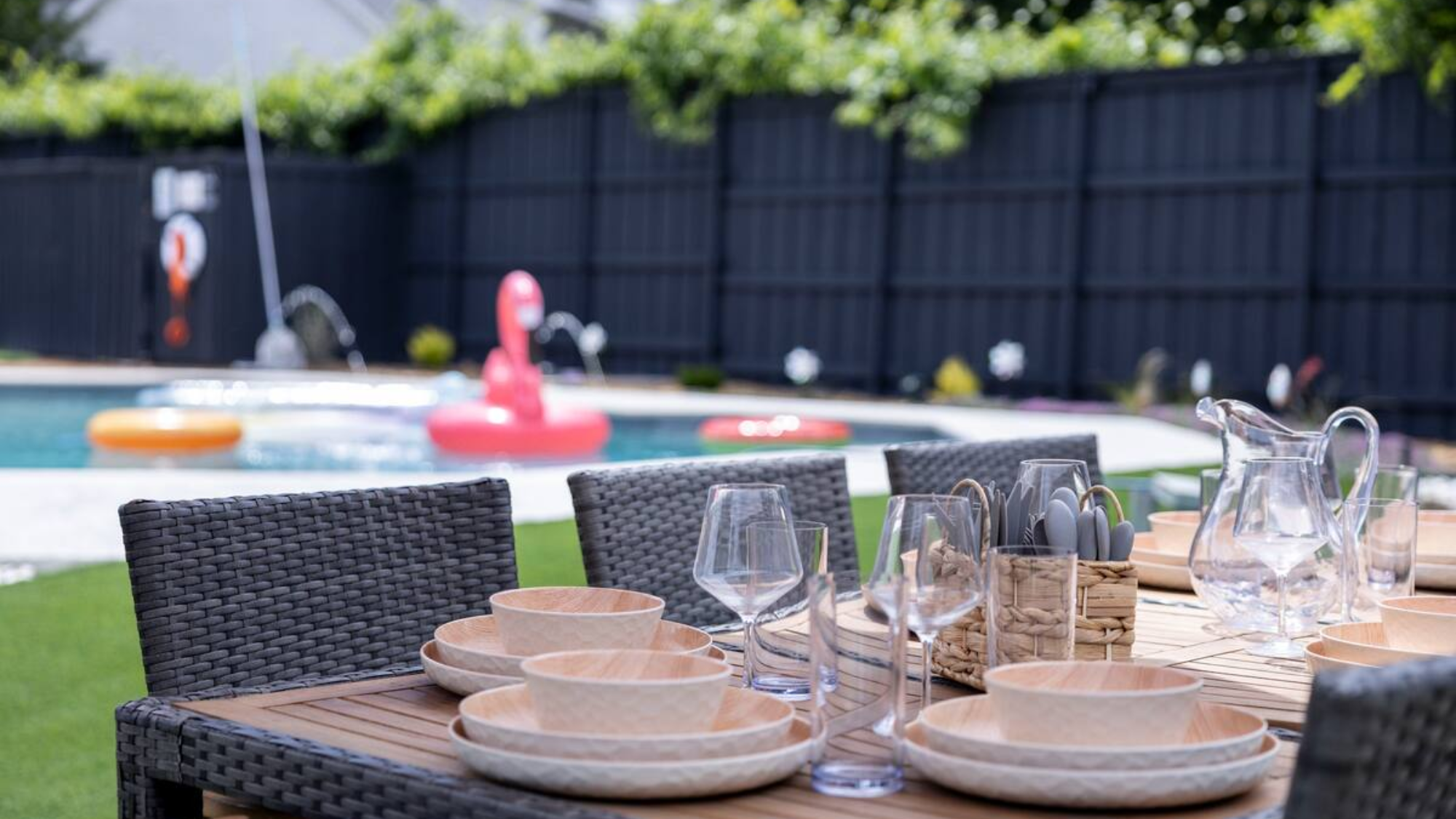 Outdoor dining setup at a family-friendly Frisco Airbnb, featuring a wooden table with woven chairs, neatly arranged plates, glasses, and utensils. In the background, a swimming pool with colorful floaties