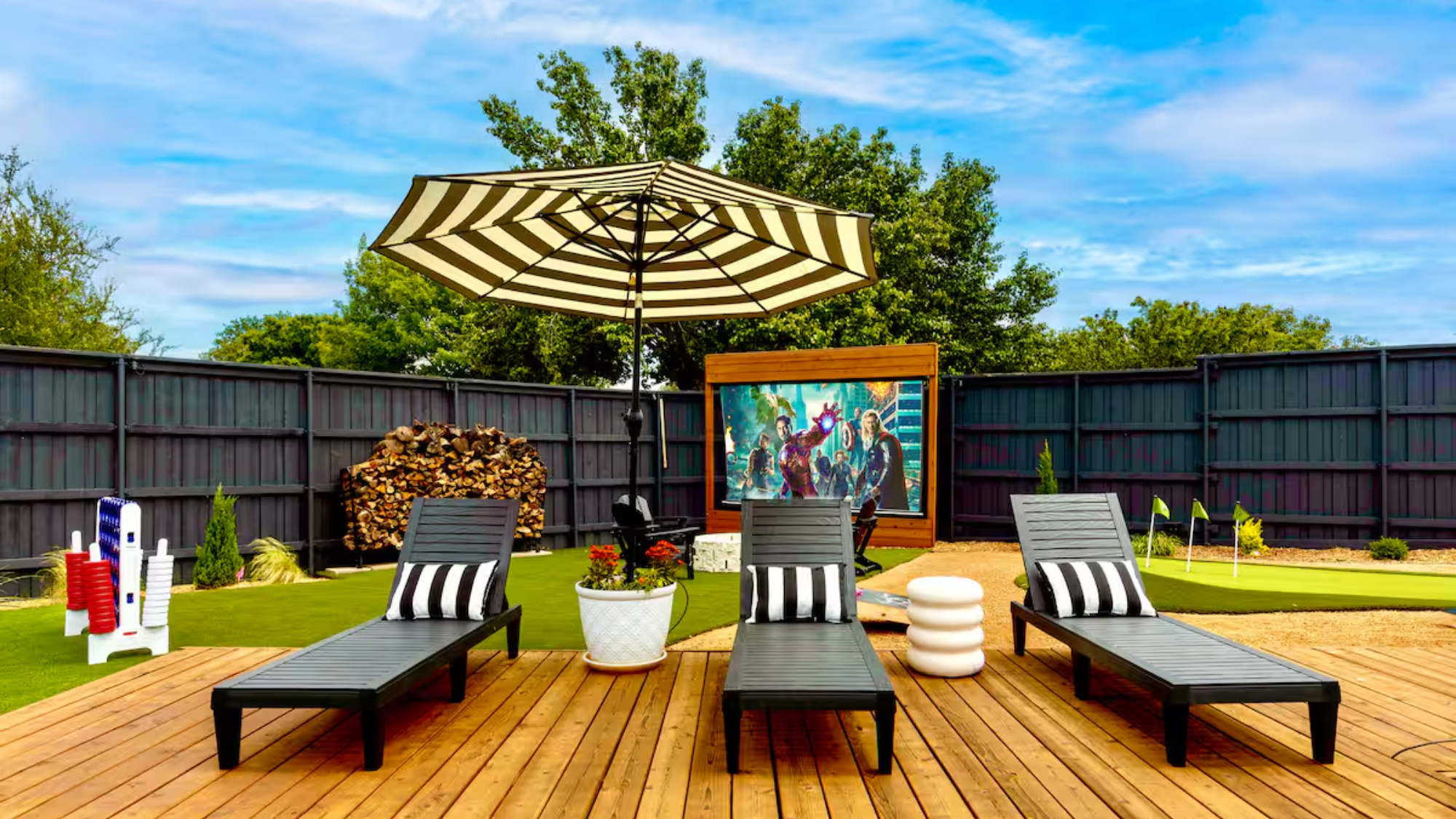 Outdoor entertainment area at a family-friendly Frisco Airbnb featuring three black lounge chairs with striped pillows on a wooden deck, a large striped umbrella, a mini-golf setup, a giant Connect Four game, and an outdoor movie screen.